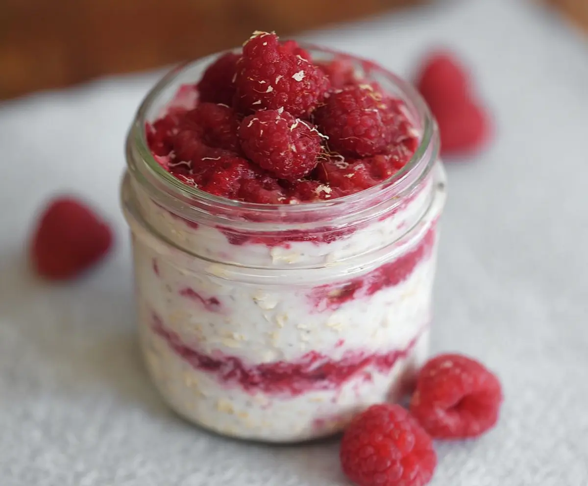 Creamy raspberry cheesecake overnight oats in a glass jar topped with fresh raspberries and mint.